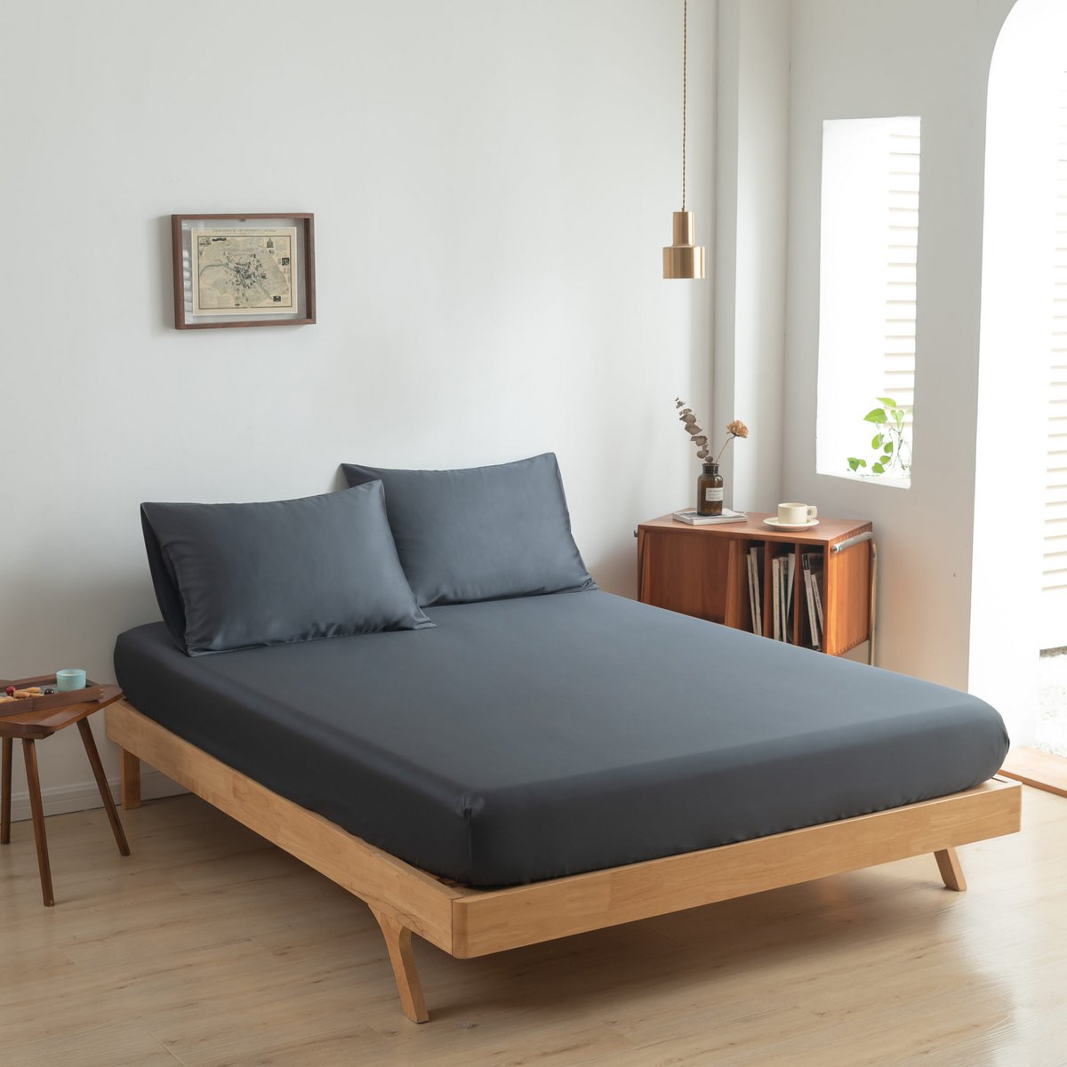 Space Blue bamboo fitted sheet on a wood-frame bed beside a window with plants.