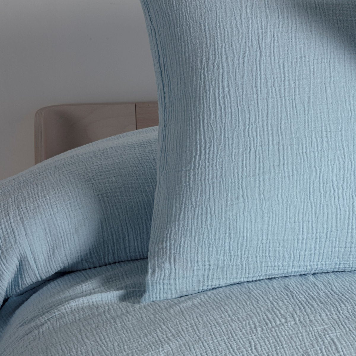 Close-up of the ice blue cotton muslin duvet cover and pillowcase showing the wrinkled texture, on a wooden bed frame