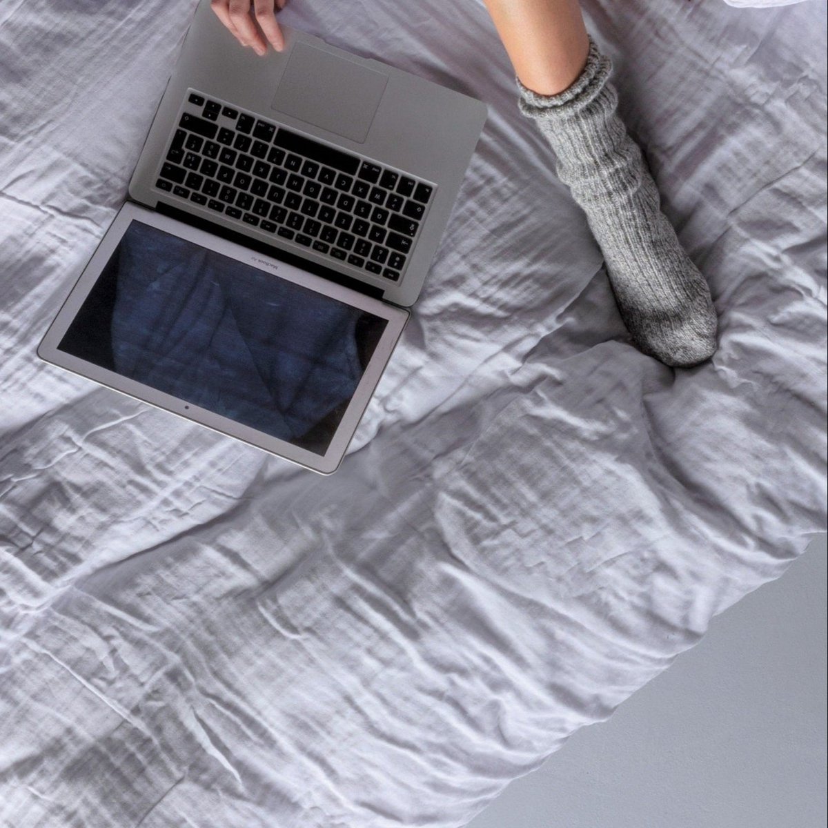Overhead view of a laptop and a grey sock resting on light grey bedding