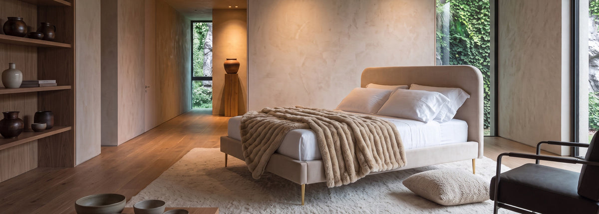Ein elegantes Polsterbett aus beigem Samt (180x200) von Maison de Cocon in einem minimalistischen, modernen Schlafzimmer. Das Bett ist mit einer gemütlichen Fell- oder Kunstfelldecke drapiert und steht auf einem weißen Plüschteppich vor einer Wand mit Strukturputz. Im Hintergrund ist ein großer Fensterbereich mit viel Grün zu sehen.