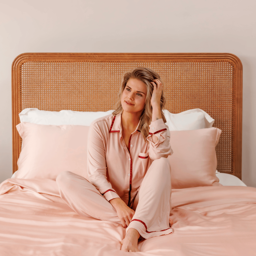 Woman sitting on a bed in front of a rattan headboard in the Powder Peach pyjama set